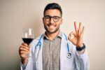 Young doctor man wearing stethoscope drinking a glass of fresh wine over isolated background doing ok sign with fingers, excellent symbol